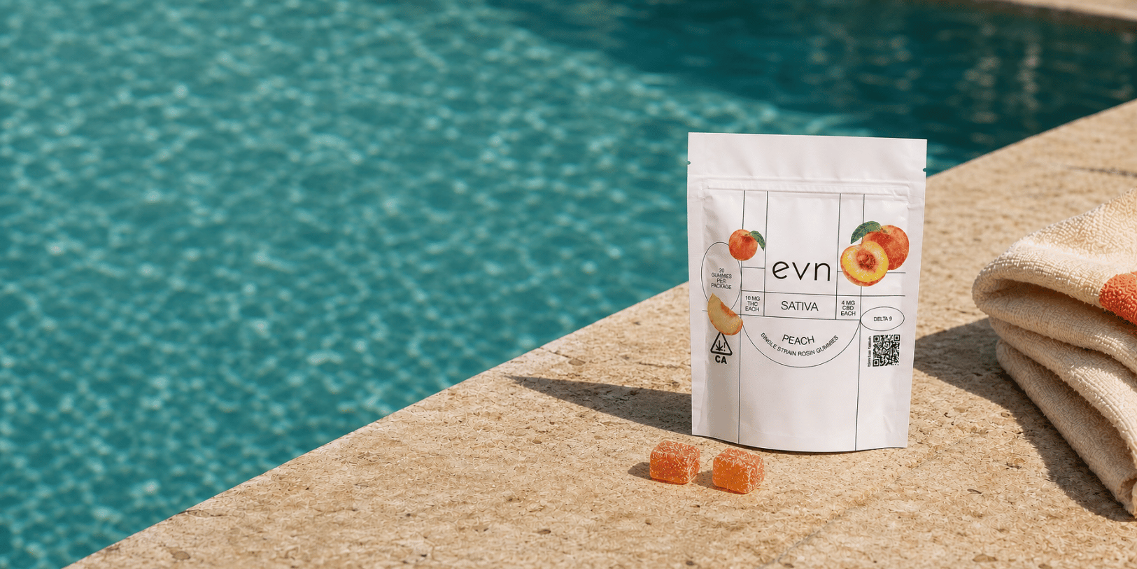 A white stand-up pouch of evn Sativa Peach Gummies sits on a stone poolside deck with a turquoise swimming pool in the background and a folded beige towel to the right.
