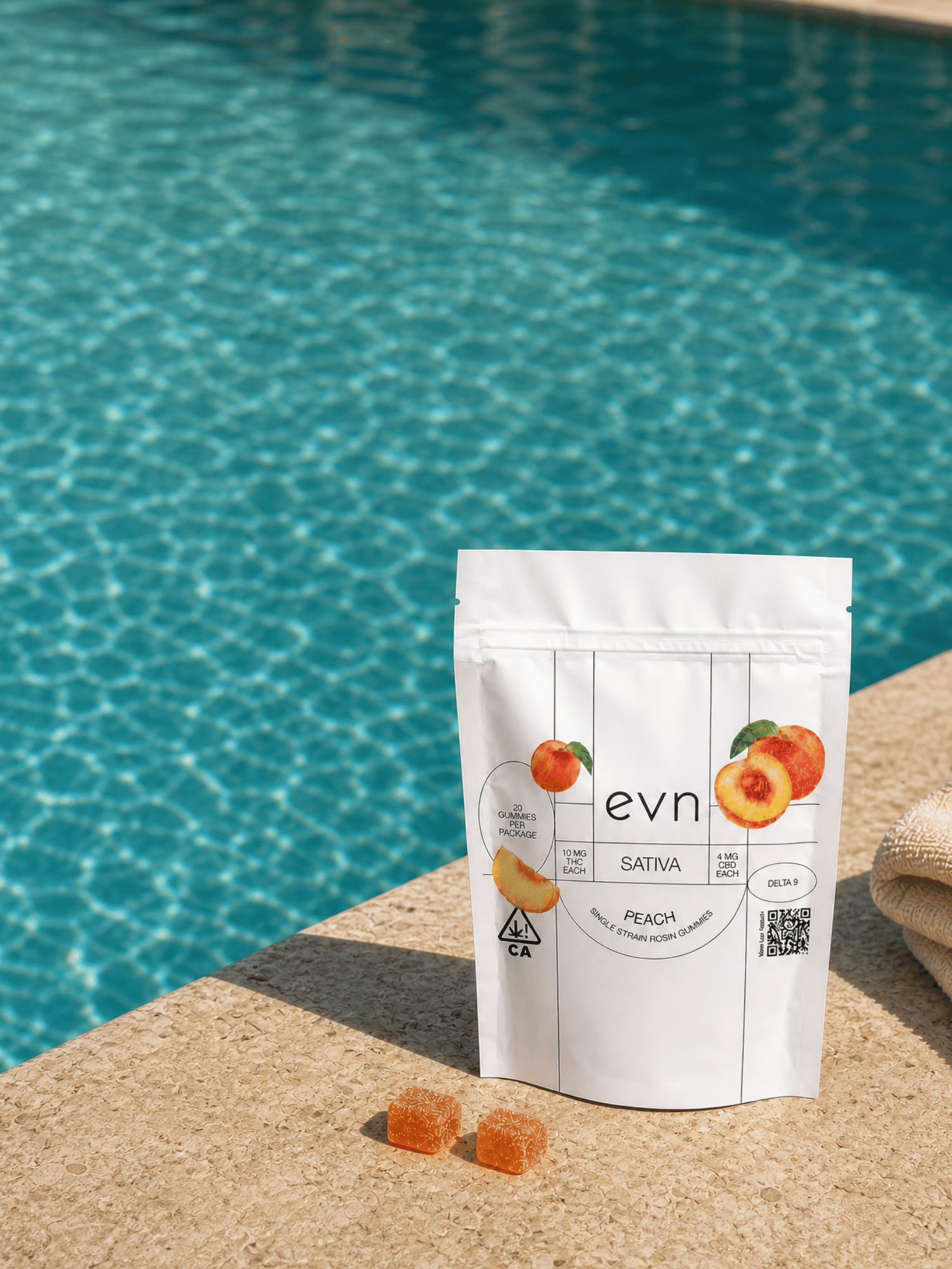 A white stand-up pouch of evn Sativa Peach gummies sits on a beige stone poolside surface with the blue swimming pool in the background. Two orange gummies are placed in front of the pouch.
