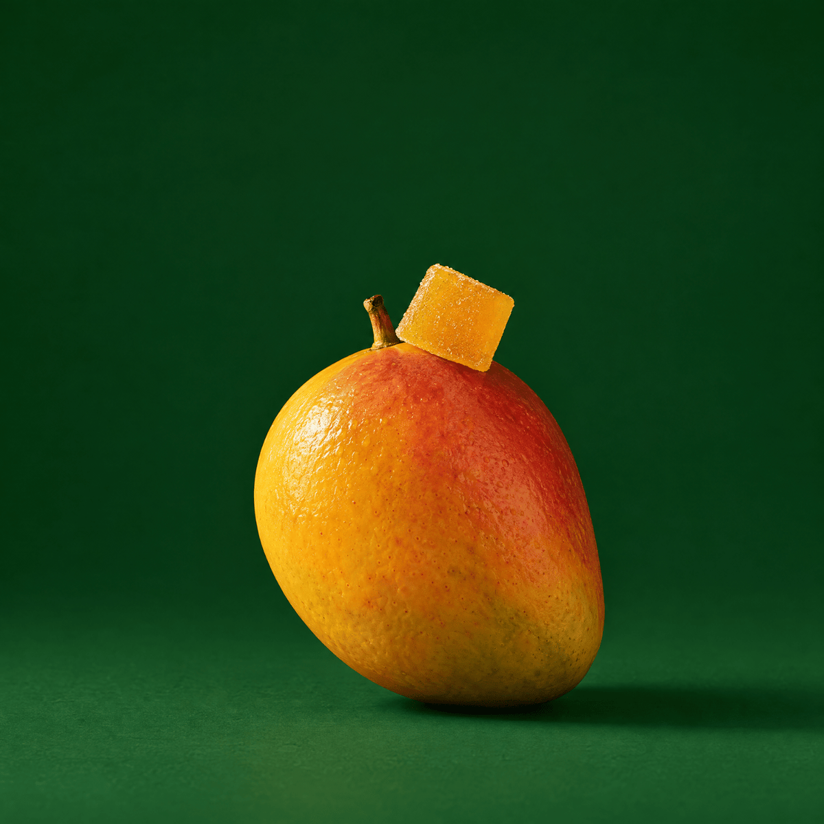A ripe mango with a small piece of orange fruit resting on top of it, photographed against a solid dark green background.