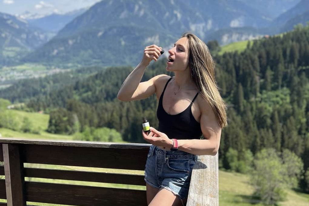 A lady consuming CBD oil standing on a wooden balcony backdropped by mountains and bushes