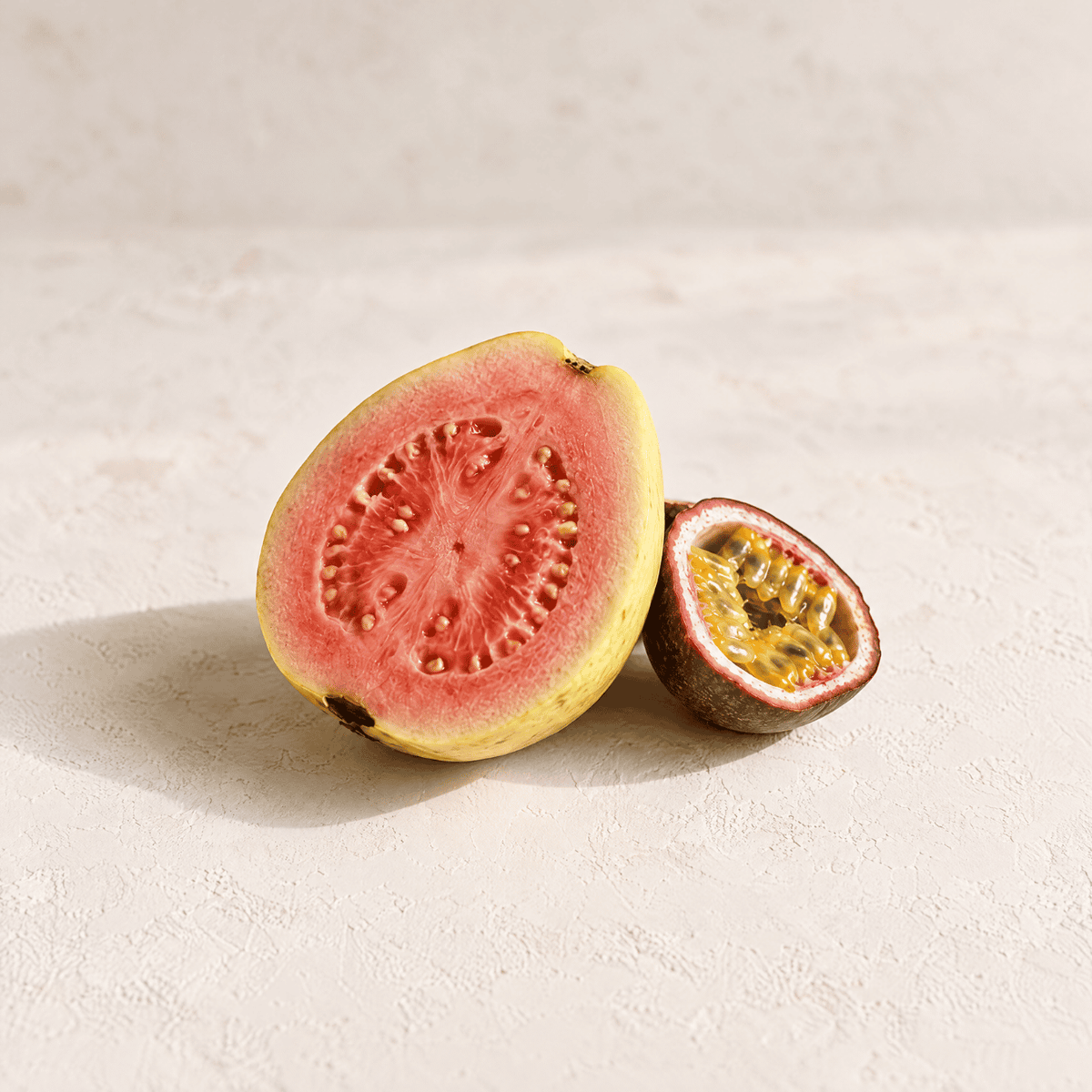 A sliced guava fruit showing pink-red flesh and yellow rind, with a smaller cut half beside it on a light textured surface.