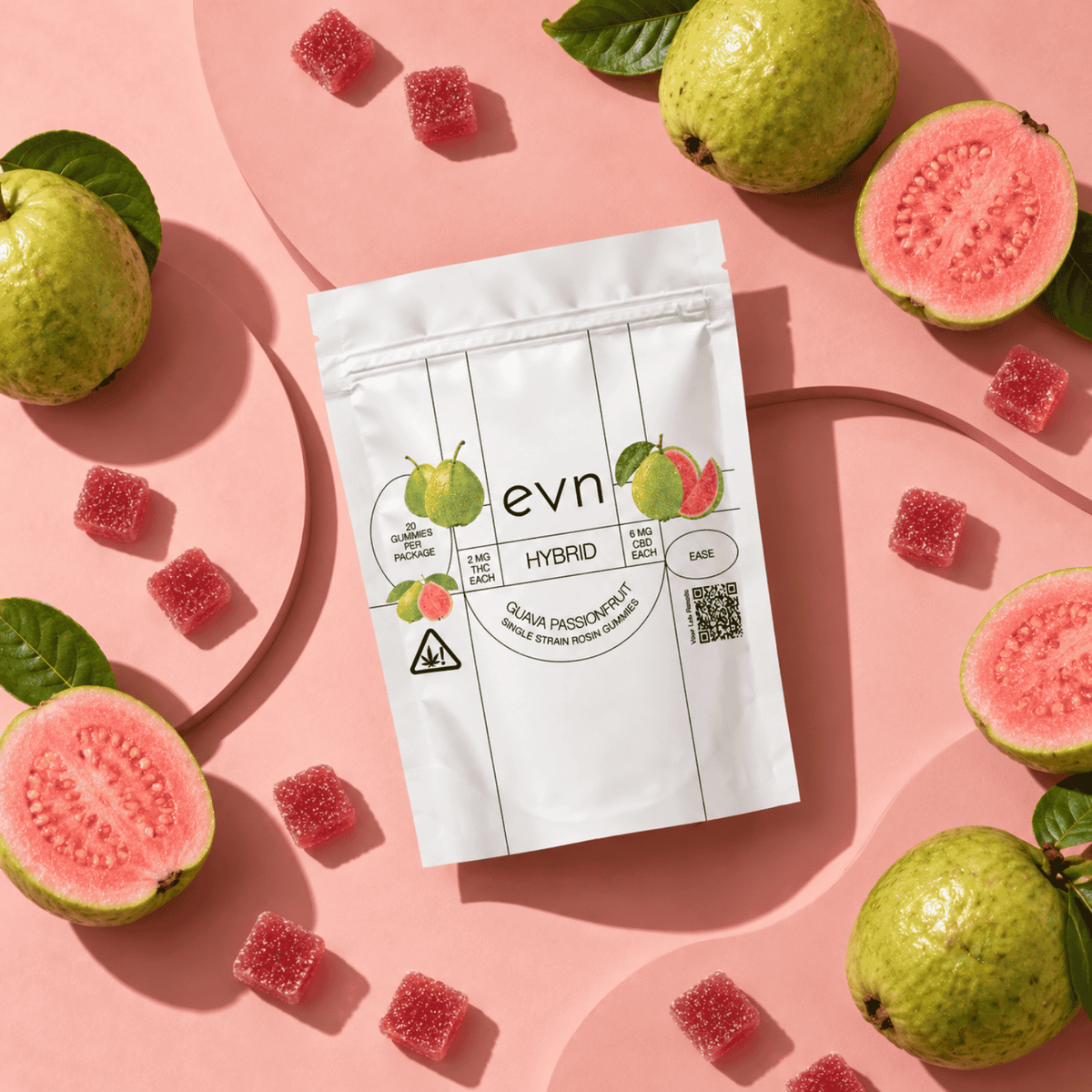 A white stand-up pouch labeled 'evn HYBRID' is arranged on a pink surface with guava fruits and pink sugar cubes around it. The pouch has fruit illustrations and a QR code, and the scene is styled as a clean flat lay.